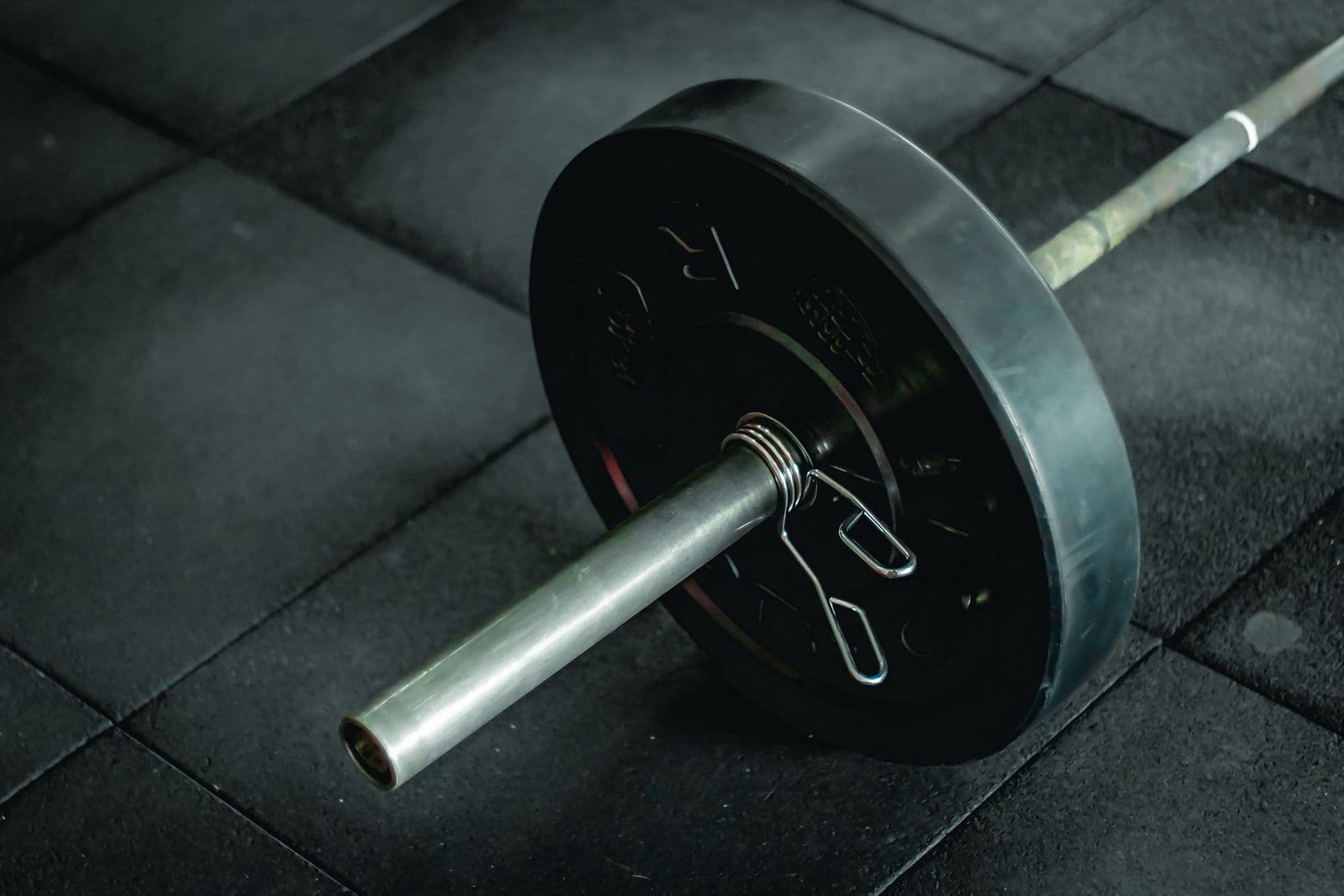 A loaded barbell resting on dark rubber gym flooring under hard light