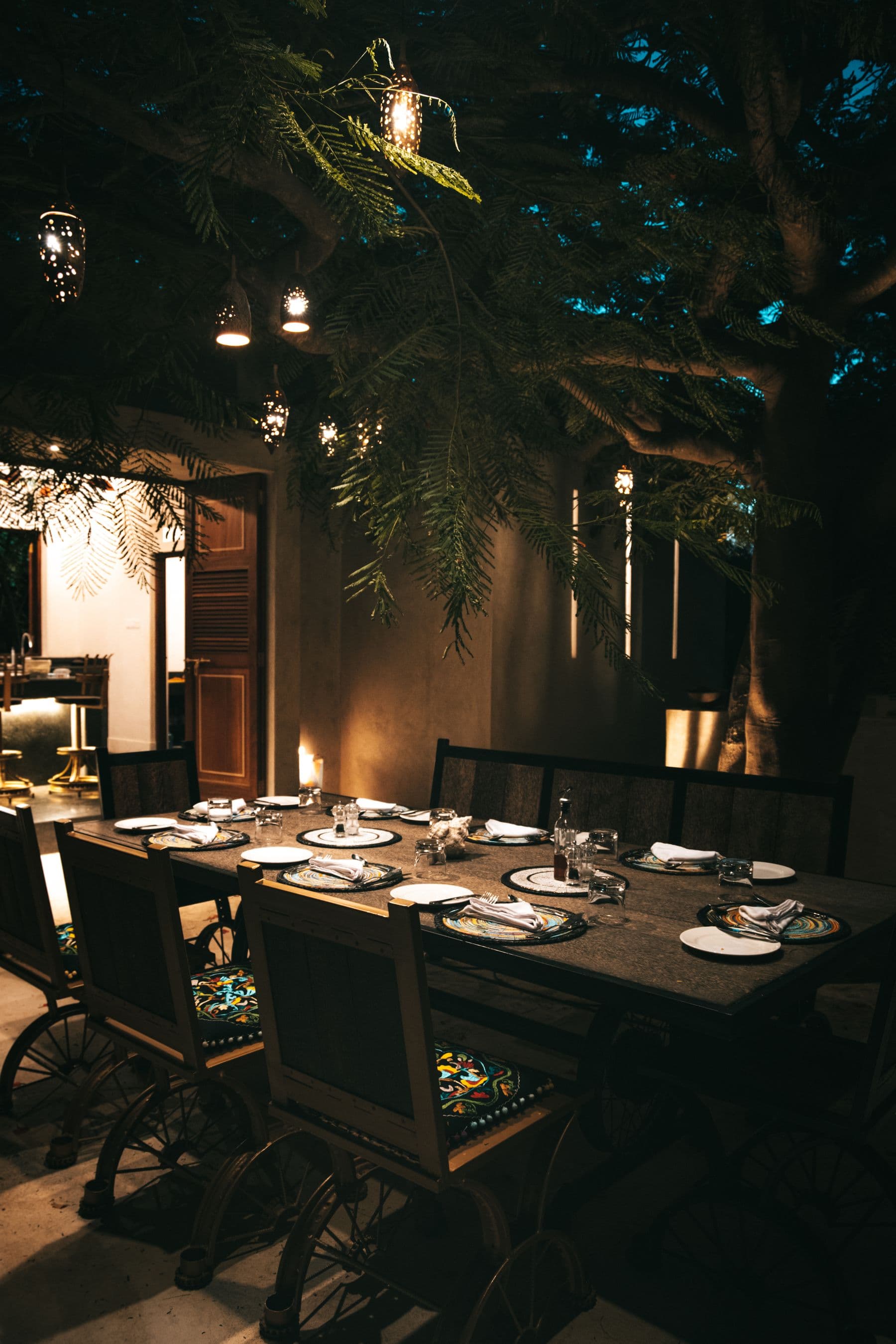 A set dinner table beneath a large tree hung with glowing lanterns at dusk